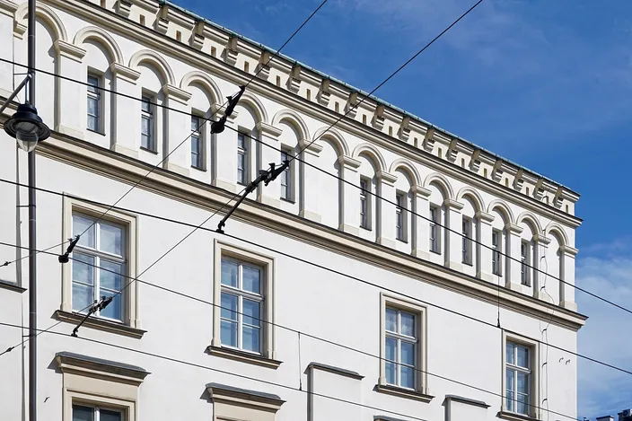 The upper two stories of a white Kazimierz facade under a deep blue sky, a row of small blind arches running below the cornice and tall shuttered windows beneath, with tram wires and a streetlamp crossing the frame.