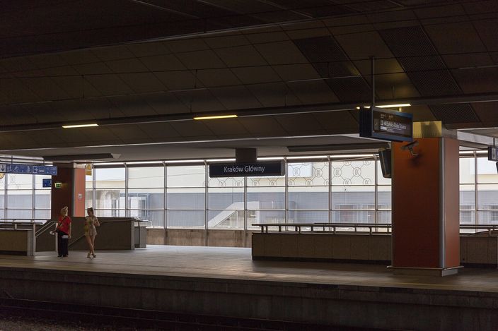 A platform at Kraków Główny station: a long empty concourse under a pale modern roof, with departure screens, a bench and two travellers waiting beside the track.
