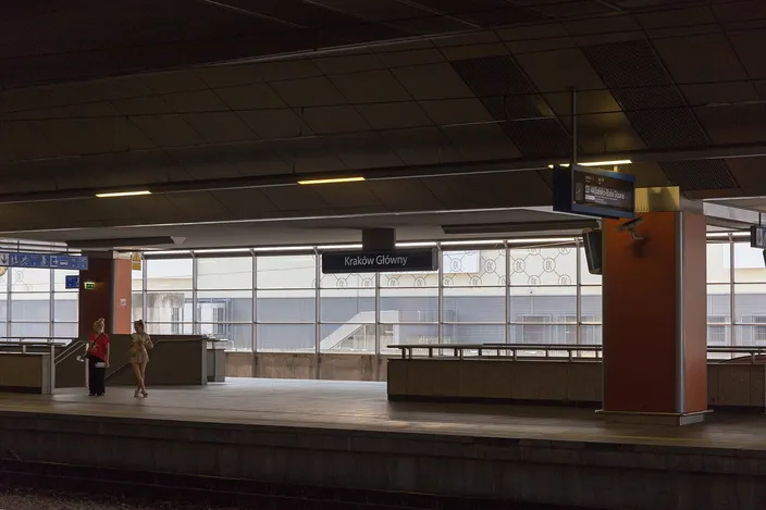 A platform at Kraków Główny station: a long empty concourse under a pale modern roof, with departure screens, a bench and two travelers waiting beside the track.