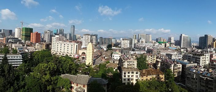 Kunming's downtown from above on a clear day: mid-rise apartment blocks and tree-lined streets running back to a line of glass towers under a blue sky with cumulus.