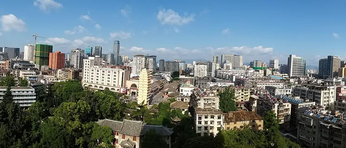Kunming's downtown from above on a clear day: mid-rise apartment blocks and tree-lined streets running back to a line of glass towers under a blue sky with cumulus.