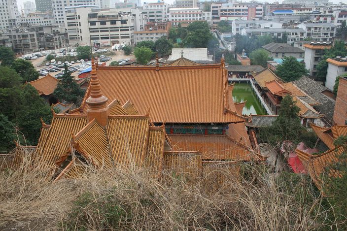 The tiered ochre-tiled roofs of a large Chinese Buddhist temple seen from the wooded slope above it, with courtyards and a small pagoda beside it and city buildings rising immediately behind.