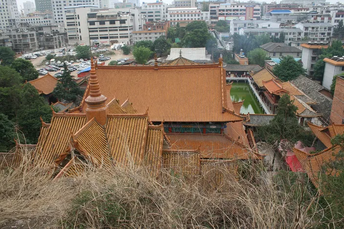 The tiered ochre-tiled roofs of a large Chinese Buddhist temple seen from the wooded slope above it, with courtyards and a small pagoda beside it and city buildings rising immediately behind.