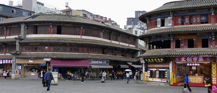 A paved public square in an old Chinese quarter, flanked by two large two-storey buildings with curved tiled eaves and timber galleries above ground-floor shopfronts, people walking across it and modern apartment blocks rising behind.