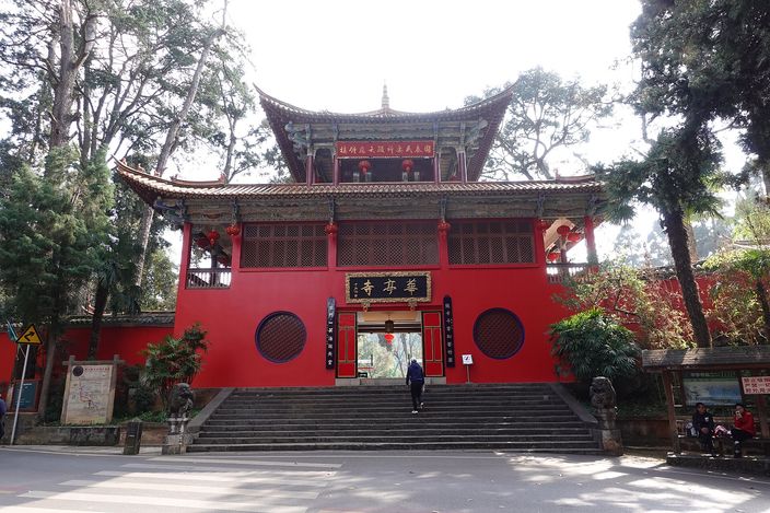 A tall red-walled Chinese temple gatehouse with a dark tiled double roof and a gilt-lettered name board, reached by a broad flight of stone steps, standing among tall pines with a single visitor climbing towards the open door.