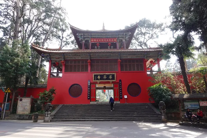 A tall red-walled Chinese temple gatehouse with a dark tiled double roof and a gilt-lettered name board, reached by a broad flight of stone steps, standing among tall pines with a single visitor climbing towards the open door.