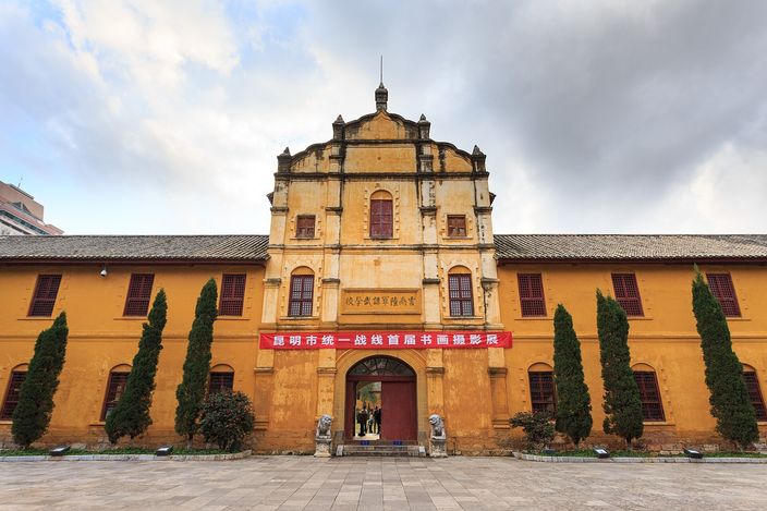 The ochre two-storey range of an early twentieth-century military academy seen across its empty paved courtyard: rows of shuttered windows, a raised central bay, a red banner over the arched doorway and a stone lion on either side of it.