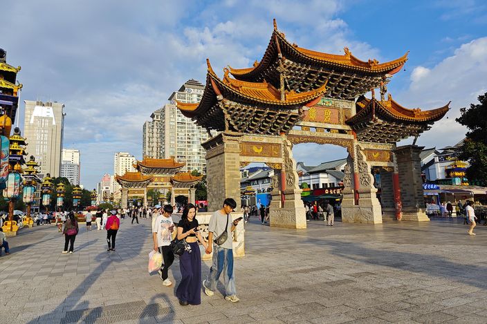 The Golden Horse and Jade Rooster archways in Jinbi Square in low evening sun, people crossing the paved square in front of them and apartment towers rising behind.