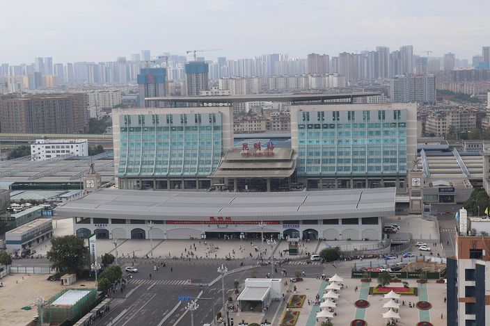 Kunming Railway Station from above: the long low station hall with its two green-glass wings, the forecourt and bus bays in front, and the city's apartment blocks receding into haze behind.