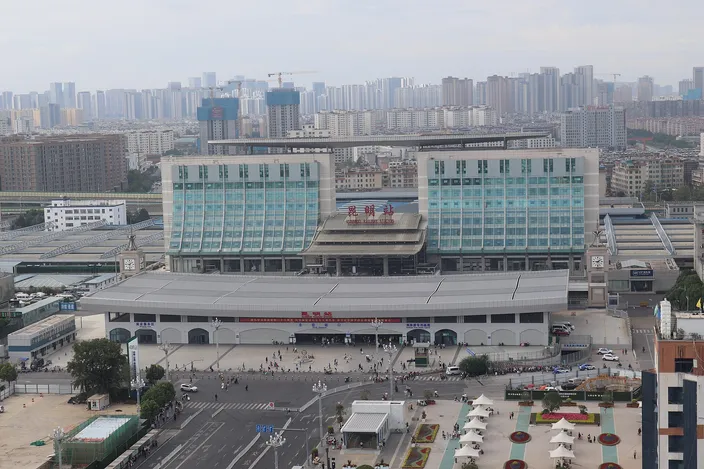 Kunming Railway Station from above: the long low station hall with its two green-glass wings, the forecourt and bus bays in front, and the city's apartment blocks receding into haze behind.