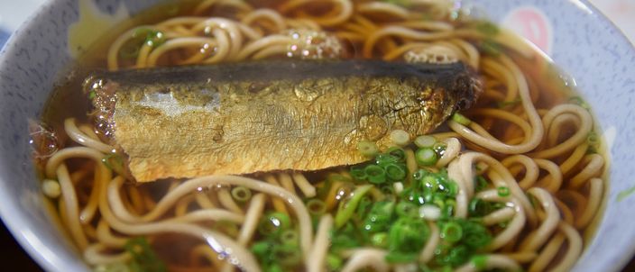 A bowl of soba in pale broth with a whole simmered herring laid across the noodles and chopped spring onion