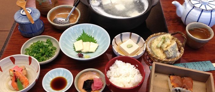 A Kyoto yudofu set: an earthenware pot of soft tofu blocks simmering in clear kelp broth, set on a tray with dipping sauce, condiments, rice and side dishes.