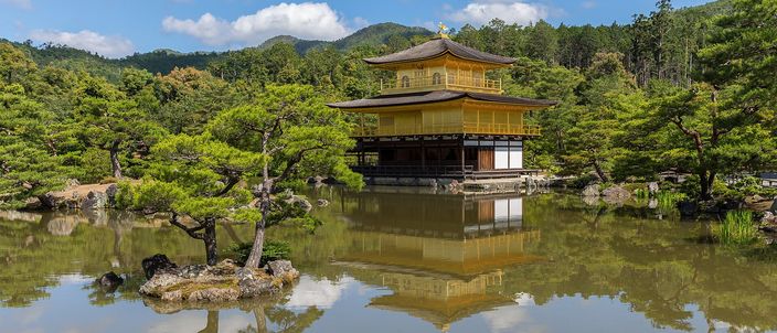 The gold-leafed Kinkaku-ji pavilion on the far bank of its pond, mirrored in still water, framed by clipped pines and wooded hills under a blue sky