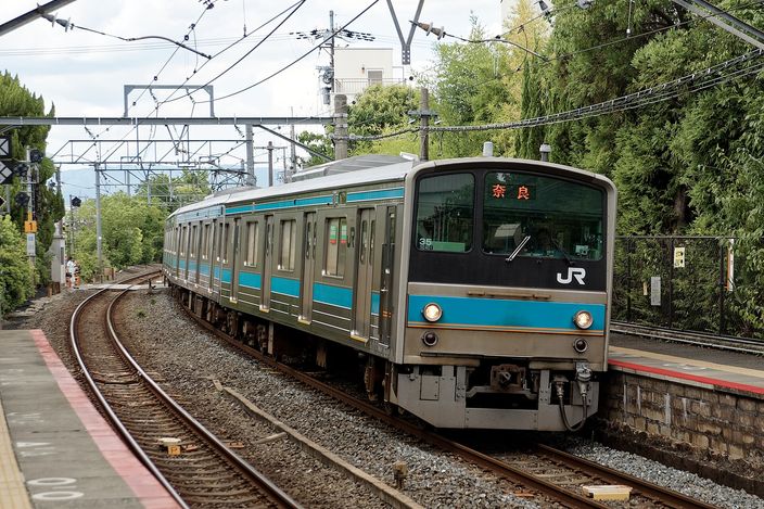 A JR West commuter train in white with a blue stripe entering Inari Station on a curve, trees and overhead wires around it and an empty platform edge in the foreground