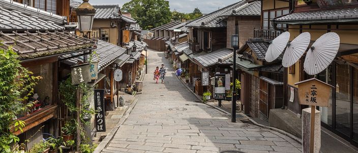 A stone-paved lane in Kyoto climbing between two-storey wooden buildings with tiled roofs, paper parasols hung on a wall to the right and a few people in yukata walking up the slope in the distance