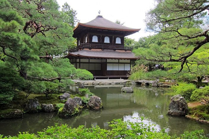 A two-storey temple pavilion of plain dark timber with a curved shingled roof, standing behind a pond with mossy rocks and clipped pines around it
