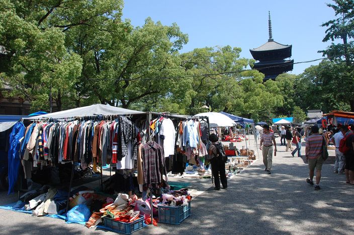 Open-air stalls of second-hand clothes and bric-a-brac spread under trees on the gravel of a temple courtyard, with a dark five-storey wooden pagoda rising behind them against a blue sky