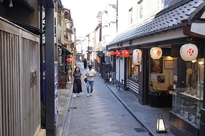 A narrow Kyoto alley at dusk between wooden machiya frontages, red and white paper lanterns hung outside restaurants, two people walking down the middle