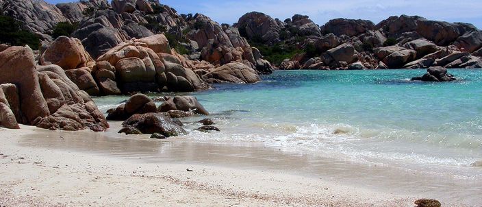 A small cove on Caprera: coarse pale sand in the foreground, rounded pink-grey granite boulders enclosing it on the left and across the water, and the sea shading from clear turquoise at the shore to deep blue beyond, under a bright sky.