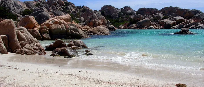 A small cove on Caprera: coarse pale sand in the foreground, rounded pink-gray granite boulders enclosing it on the left and across the water, and the sea shading from clear turquoise at the shore to deep blue beyond, under a bright sky.