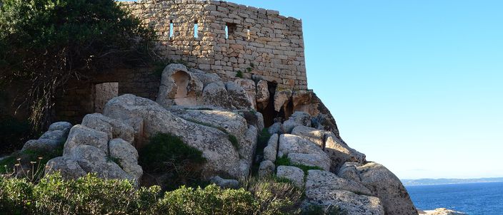 The ruined stone wall of the Forte di Punta Tegge standing on a shelf of rounded granite boulders at the island's south-west tip, low scrub in the foreground and the sea and the Sardinian coast beyond, with a strip of shallow turquoise water at the right-hand edge.