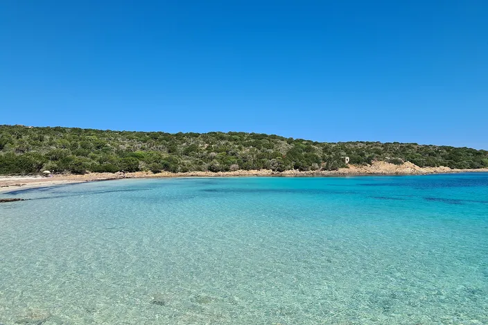 A crescent cove of pale sand meets clear water shading from pale green to turquoise, backed by a low headland of green Mediterranean scrub under a bright blue sky; no wreck or people in view.