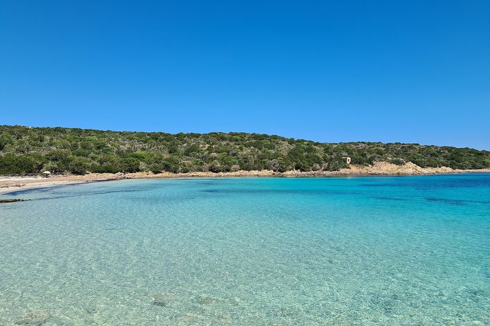 A crescent cove of pale sand meets clear water shading from pale green to turquoise, backed by a low headland of green Mediterranean scrub under a bright blue sky; no wreck or people in view.