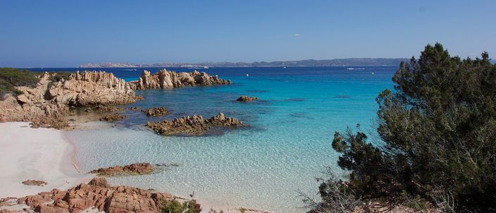 A granite cove on Budelli seen from the rocks above: pink-white sand curving around a shallow turquoise bay, red granite boulders and Mediterranean maquis in the foreground, hazy hills across the water beyond.