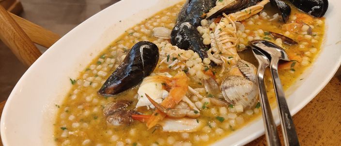 A restaurant plate of fregola ai frutti di mare — pearl-shaped semolina pasta in a saffron-orange seafood broth, with mussels, clams and prawns.