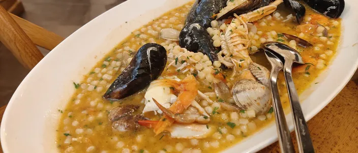 A restaurant plate of fregola ai frutti di mare — pearl-shaped semolina pasta in a saffron-orange seafood broth, with mussels, clams and prawns.