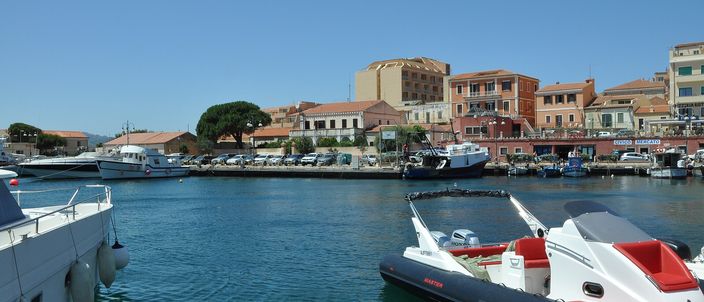Cala Gavetta harbour front in La Maddalena: colourful granite-built townhouses and a church dome behind moored fishing boats and RIBs in the foreground water, under a clear sky.