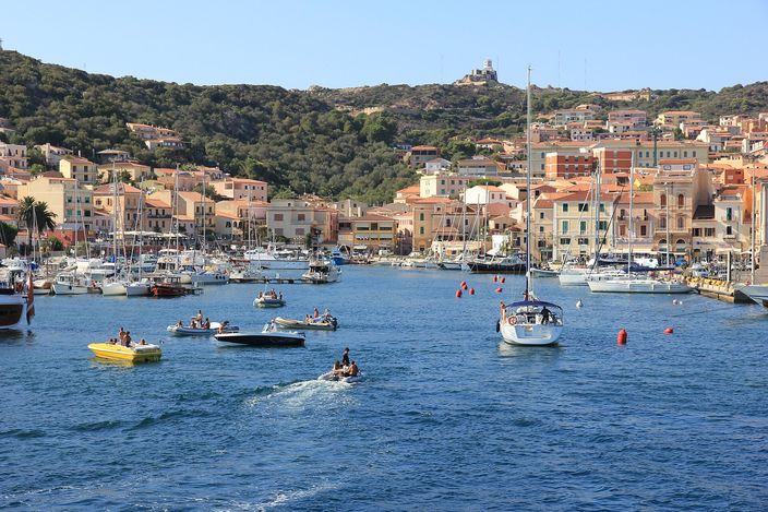 A wide view of La Maddalena's harbour of Cala Gavetta from the water, with sailboats moored in front and the pastel-coloured town rising up the hillside behind, a tower visible on the skyline.