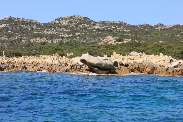A large, smoothly weathered pink granite boulder rising out of turquoise water off Budelli, with a low ridge of similar wind-sculpted granite rocks beside it and green macchia-covered hills behind.