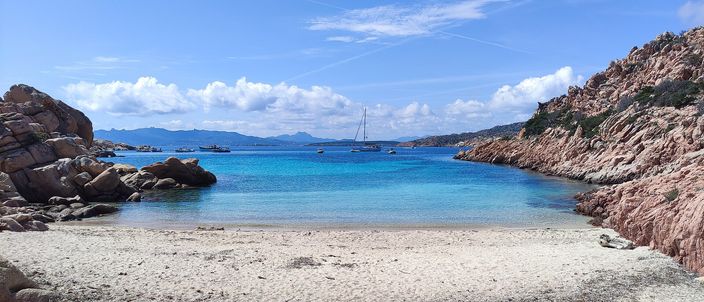 A small cove on Caprera: pale sand in the foreground, rounded pink-orange granite boulders framing turquoise water, and a single sailboat anchored in the bay under a blue sky.
