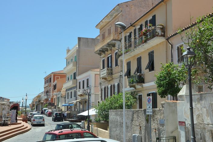 Piazza des Geneys in the old town: a whitewashed, red-brick-trimmed townhouse with a balcony at the head of a pedestrian square, café tables under awnings and a few strollers in the midday sun.
