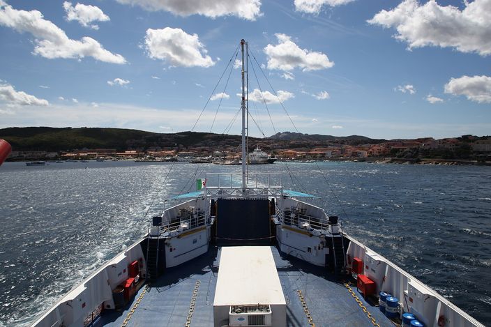 View forward from the bow deck of a car ferry mid-crossing, the mast and funnel of the ship in frame, La Maddalena's waterfront and hills visible ahead across the strait.