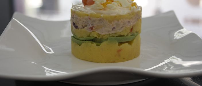 A single serving of causa limeña on a white ruffled-edge plate: a tower of yellow mashed potato rests on a slice of avocado, topped with a dollop of white dressing and diced garnish, against a softly blurred kitchen background.