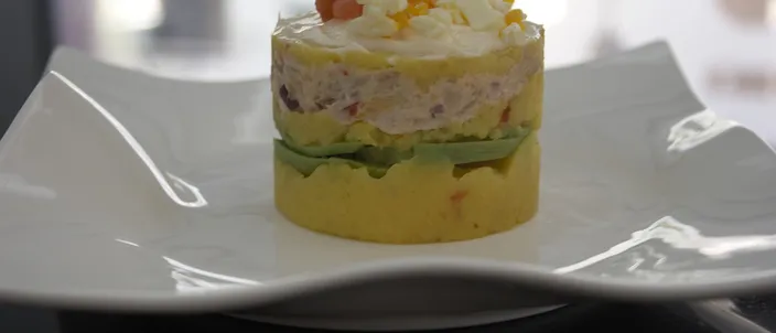 A single serving of causa limeña on a white ruffled-edge plate: a tower of yellow mashed potato rests on a slice of avocado, topped with a dollop of white dressing and diced garnish, against a softly blurred kitchen background.