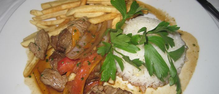 A plate of lomo saltado in Lima: strips of seared beef with onion, tomato and red pepper, a pile of fried potato strips, and a mound of white rice with parsley on the side.
