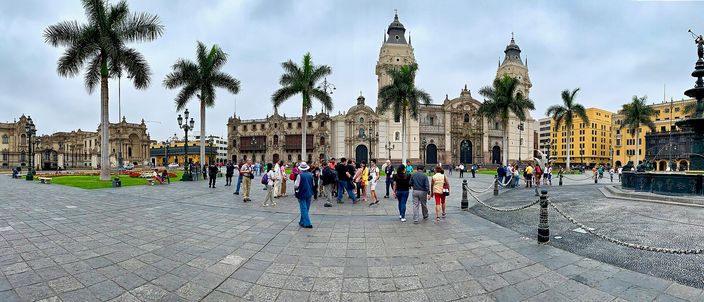 A wide panorama of Lima's Plaza Mayor at dusk, with the cathedral's twin towers, the yellow colonnaded Archbishop's Palace, palm trees, a central fountain and tourists crossing the square.