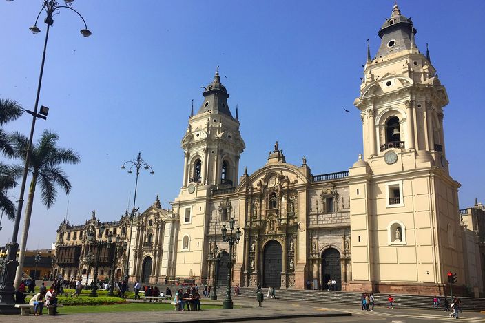 Lima Cathedral's twin baroque bell towers and ochre stone facade rise over Plaza Mayor under a clear blue sky, with palm trees, a lamp post and pedestrians in the foreground.
