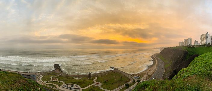 A panoramic sunset view along the Miraflores clifftop: a golden-orange sky over the Pacific, the coastal road curving beneath green cliffside parks, apartment towers rising at the far end.