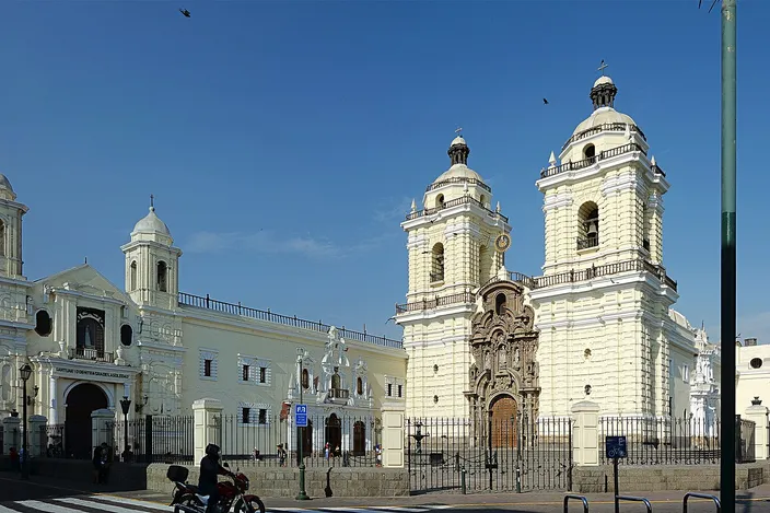The white colonial facade of the Convento de San Francisco de Asís, with its two bell towers and the adjoining church beside it, seen from the street with a traffic light and lamp post in the foreground.