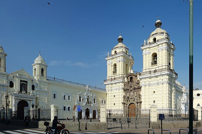 The white colonial facade of the Convento de San Francisco de Asís, with its two bell towers and the adjoining church beside it, seen from the street with a traffic light and lamp post in the foreground.