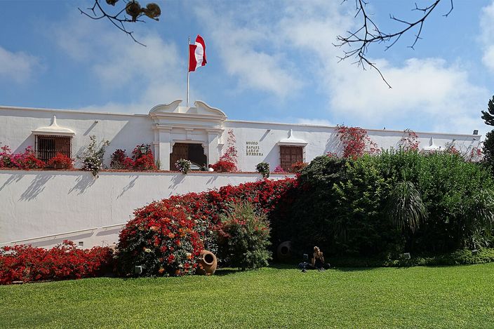 The white colonial-era building of the Museo Larco framed by a mass of red bougainvillea in bloom, the Peruvian flag flying above the entrance and a lawn in front.