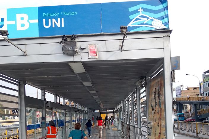 A covered elevated walkway at a Metropolitano bus station in Lima, with passengers walking along the platform under blue station signage reading "Estación UNI".
