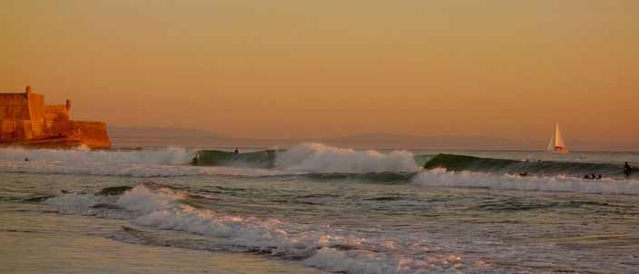 Surfers riding lines of Atlantic swell breaking across Praia de Carcavelos at sunset, with the low stone bastions of the Forte de Sao Juliao da Barra silhouetted on the headland at the left of the frame and a white sail on the horizon.