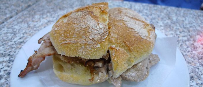A bifana on a white plate: a crusty floured Portuguese roll cut in half, filled with thin slices of pale simmered pork, on a granite café counter.