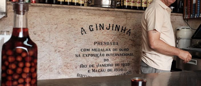 A tall glass jar of whole sour cherries steeping in deep-red ginjinha on a marble bar counter, with a small poured glass of the liqueur beside it and a bartender and rows of bottles behind.