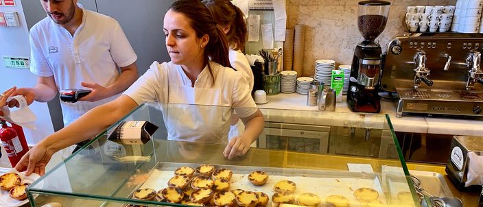 The counter of a Lisbon pastelaria: a glass case filled with rows of scorch-topped pasteis de nata, white-shirted staff plating tarts behind it, an espresso machine and stacked cups against the wall.